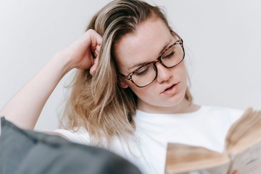 Low angle of young female in eyeglasses wearing white t shirt leaning on hand and reading book